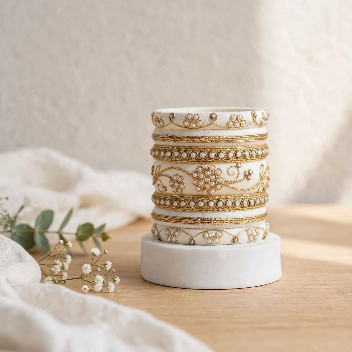 A stack of five white cylindrical candle jars with ornate gold floral and vine patterns, displayed on a white circular base on a wooden surface with soft white fabric and small flowers in the foreground.