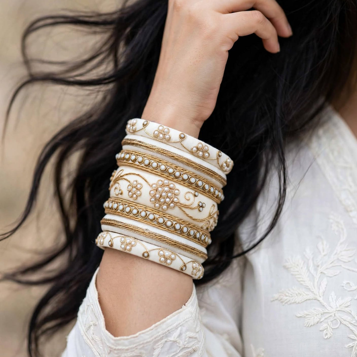 A close-up of a person wearing a stack of ornate cream and gold bangles with floral and paisley patterns, paired with a white embroidered top. The person has long, dark hair and is shown from the chest up against a softly blurred neutral background.
