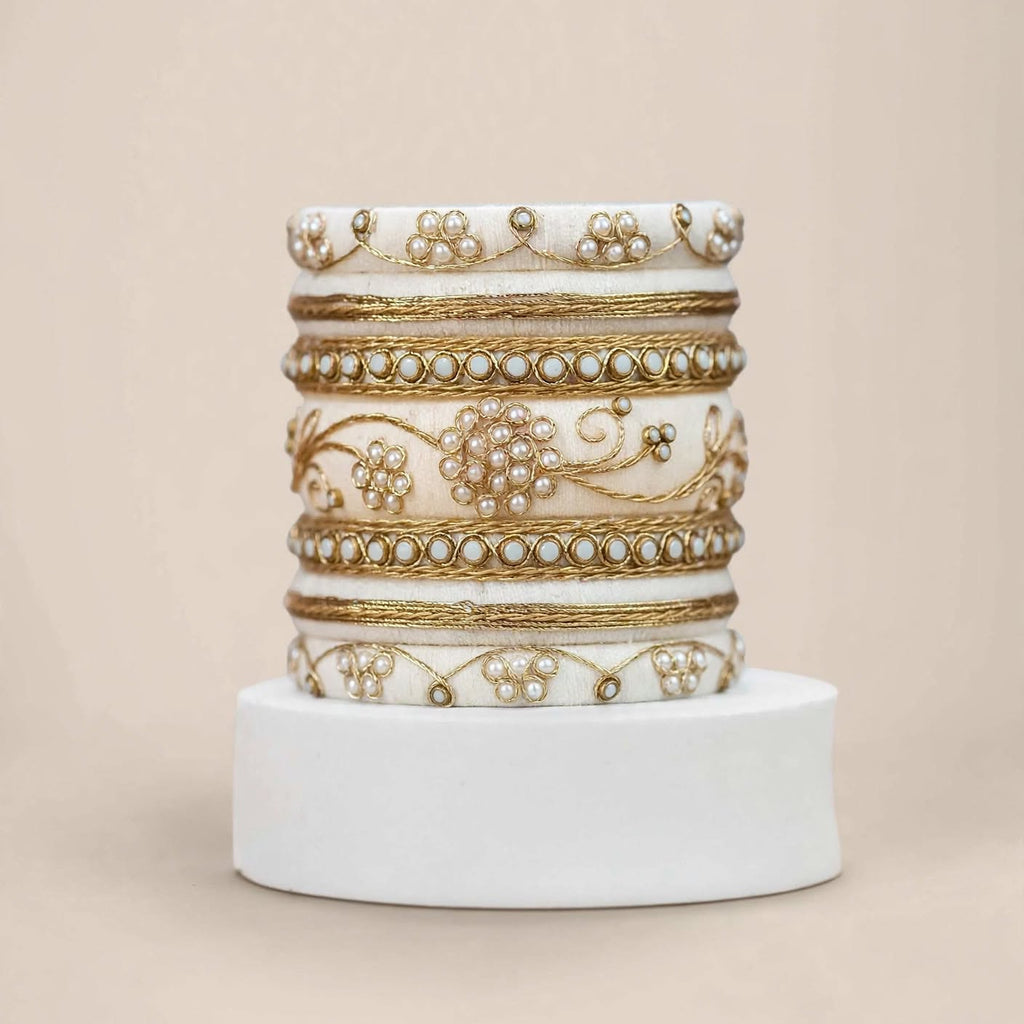 A stack of five ornate white bangles with intricate gold foil work, bead borders, and pearl-like embellishments, displayed on a white cylindrical pedestal against a solid beige background.