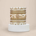 A stack of five ornate white bangles with intricate gold foil work, bead borders, and pearl-like embellishments, displayed on a white cylindrical pedestal against a solid beige background.
