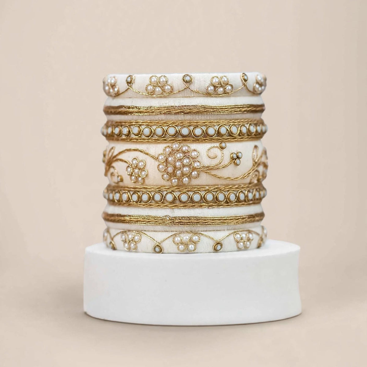 A stack of five ornate white bangles with intricate gold foil work, bead borders, and pearl-like embellishments, displayed on a white cylindrical pedestal against a solid beige background.