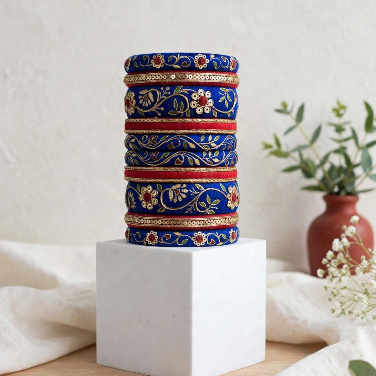 A stack of five ornate blue and gold bangles displayed on a white cube pedestal, with a neutral textured background and a small vase of greenery in the distance.