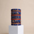 A stack of ornate blue bangles with gold floral patterns and red accents, displayed on a white cube pedestal against a beige background.