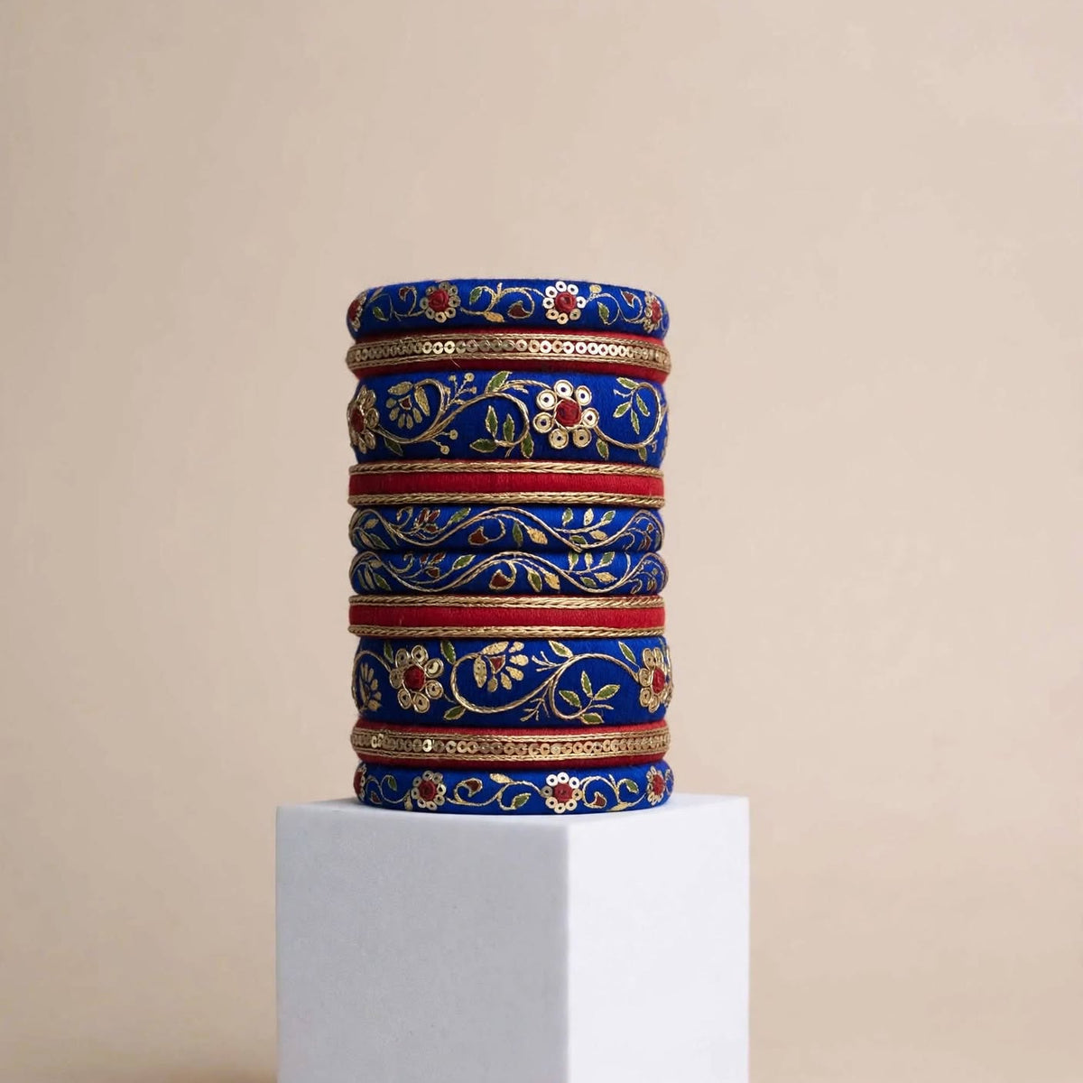 A stack of ornate blue bangles with gold floral patterns and red accents, displayed on a white cube pedestal against a beige background.