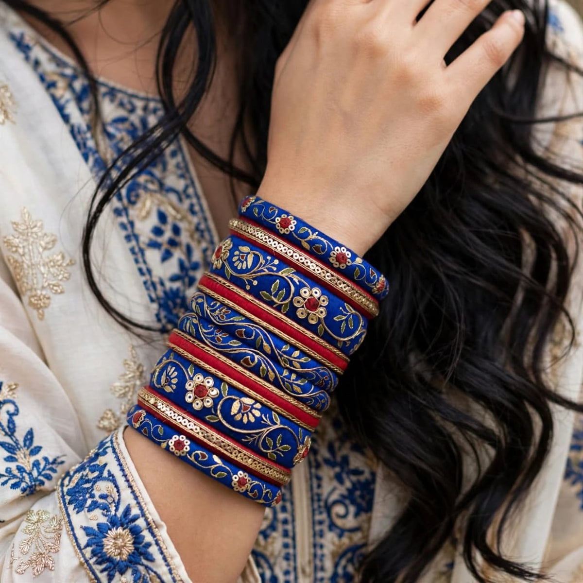 A close-up of a person wearing a stack of ornate blue bangles with red and gold floral patterns, paired with a cream embroidered kurta.