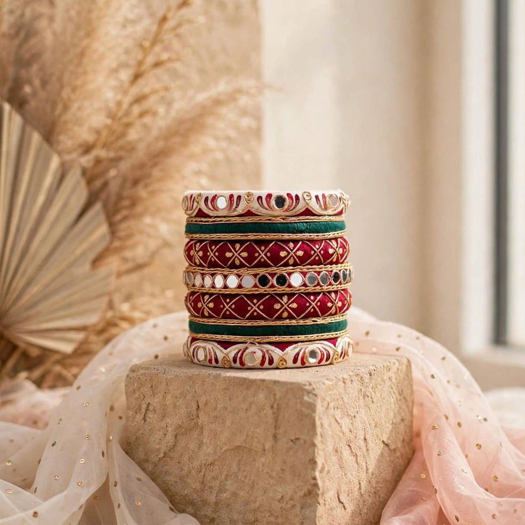 A stack of six traditional Indian bangles with intricate red, green, and gold patterns and mirror work, displayed on a stone block with soft pink and white fabric drapes in a warm, sunlit setting.