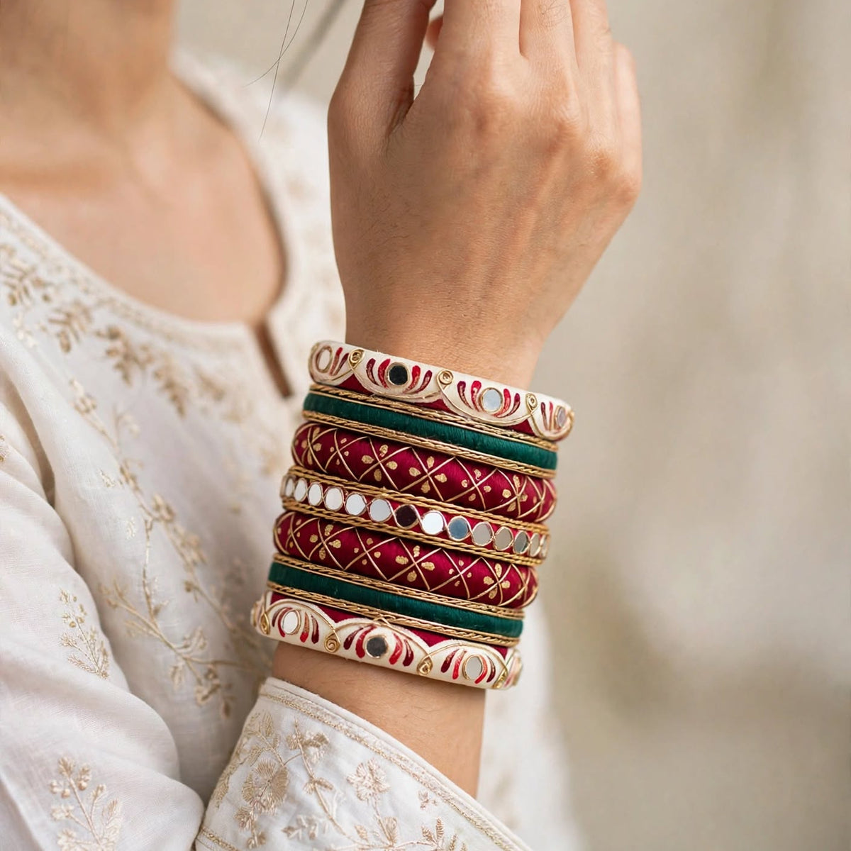 A close-up of a person wearing a stack of ornate bangles on their wrist. The bangles feature gold-plated metal borders, deep red and teal green enamel sections, and small white stone accents.
