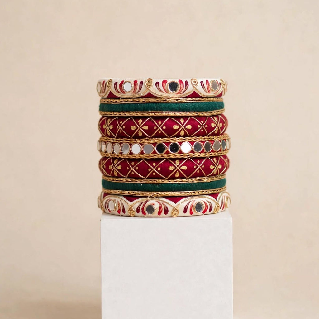 A stack of five traditional Indian Kundan bangles displayed on a white rectangular pedestal against a warm beige background. The bangles feature red enamel, gold-plated metal borders, green enamel accents, and white stone embellishments.