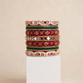 A stack of five traditional Indian Kundan bangles displayed on a white rectangular pedestal against a warm beige background. The bangles feature red enamel, gold-plated metal borders, green enamel accents, and white stone embellishments.