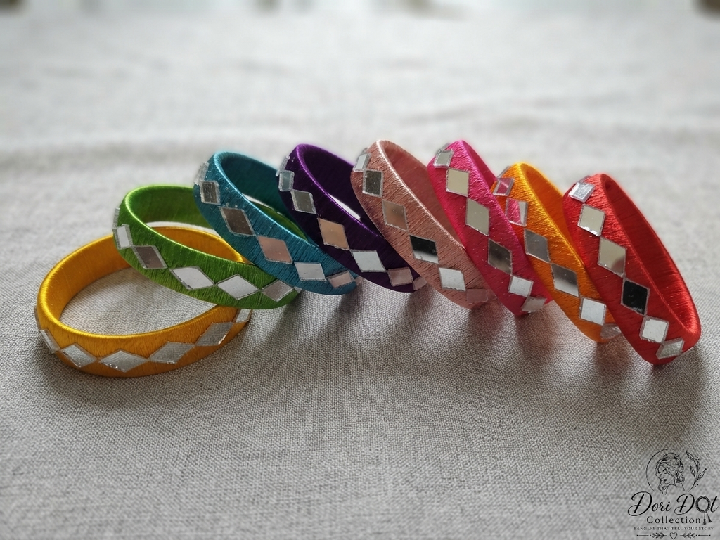 Six colorful leather bangles with mirrored silver diamond inlays, arranged in a fan shape on a textured light gray fabric background. The bangles are yellow, green, teal, purple, pink, and orange.