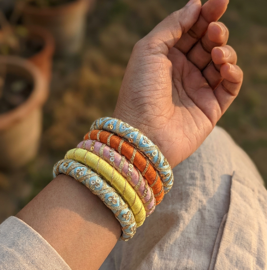 A close-up of a person's wrist wearing a stack of six woven fabric bangles in pastel colors including yellow, orange, pink, and blue, with a blurred outdoor background.