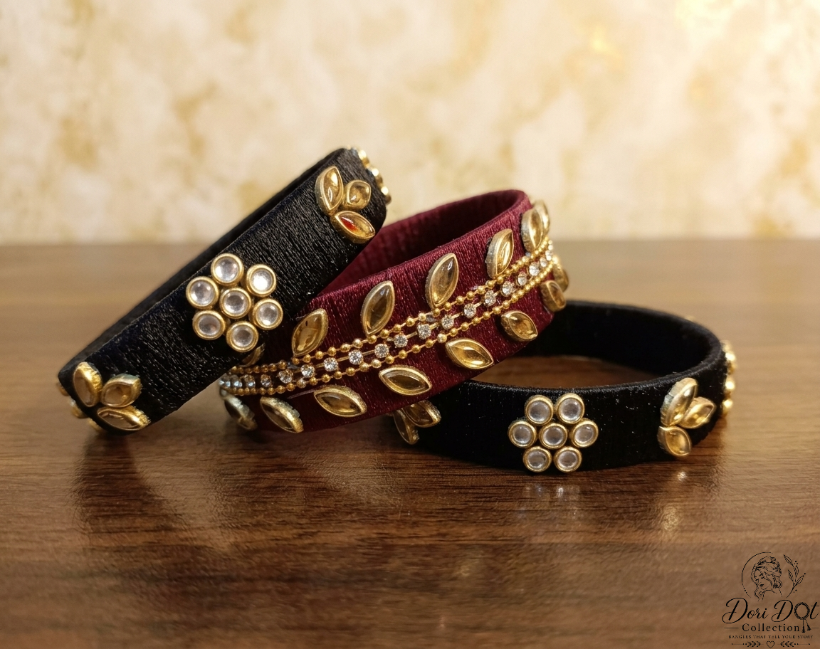 Three velvet bangles in black and deep maroon, decorated with gold-toned floral and leaf motifs and clear rhinestone flower clusters, arranged on a wooden surface.