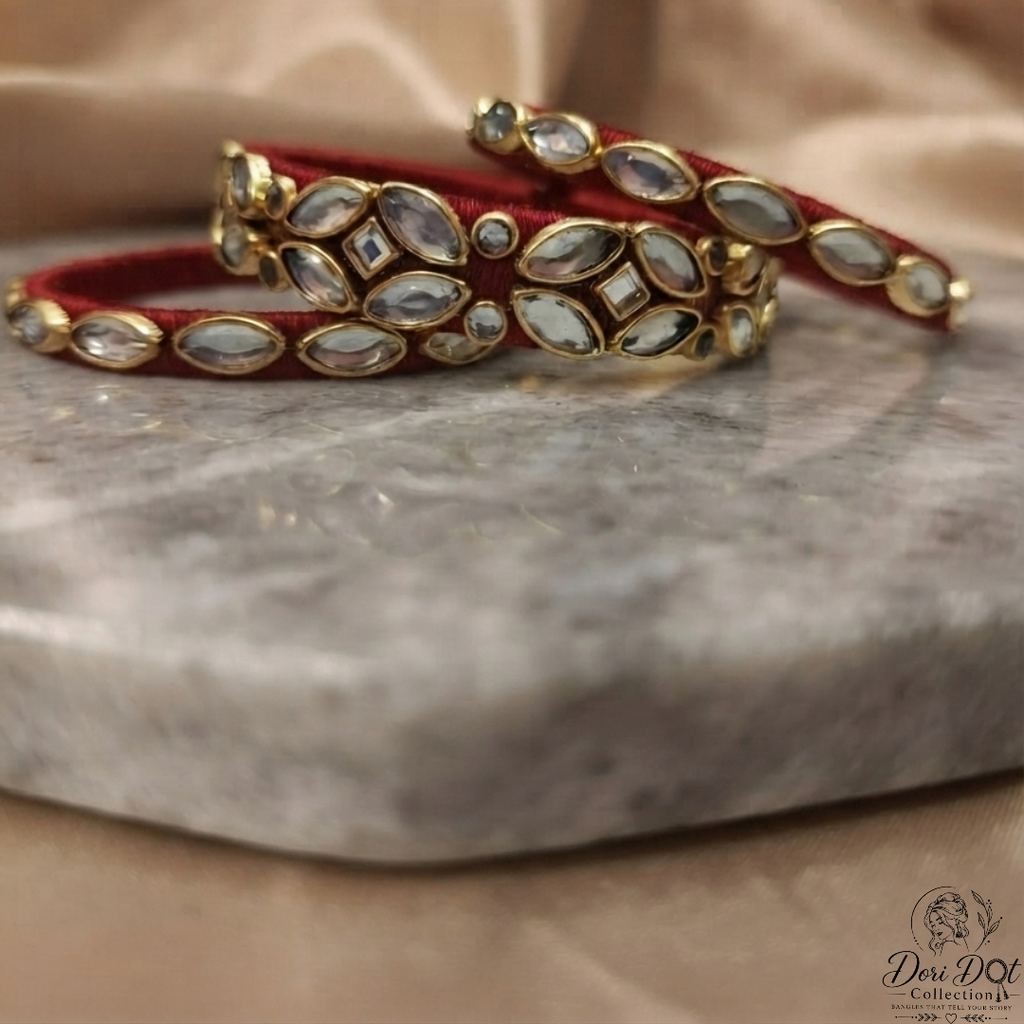 A pair of red velvet bangles with gold-plated borders and inlaid white stones, displayed on a gray marble slab with a beige satin fabric background.