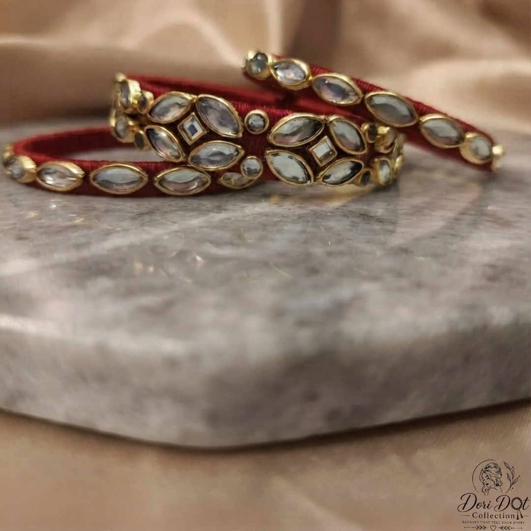 A pair of red velvet bangles with gold-plated borders and inlaid white stones, displayed on a gray marble slab with a beige satin fabric background.