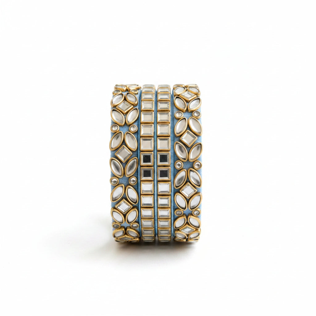 A stack of three wide gold-toned bangles with intricate floral and rectangular inlay patterns, displayed inside an open light blue jewelry box lined with white satin, surrounded by small white flowers on a white fabric background.