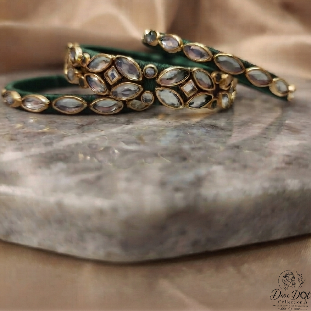 A stack of three gold-plated bangles decorated with green enamel and clear stone inlays, displayed on a gray marble slab with a beige satin fabric background.