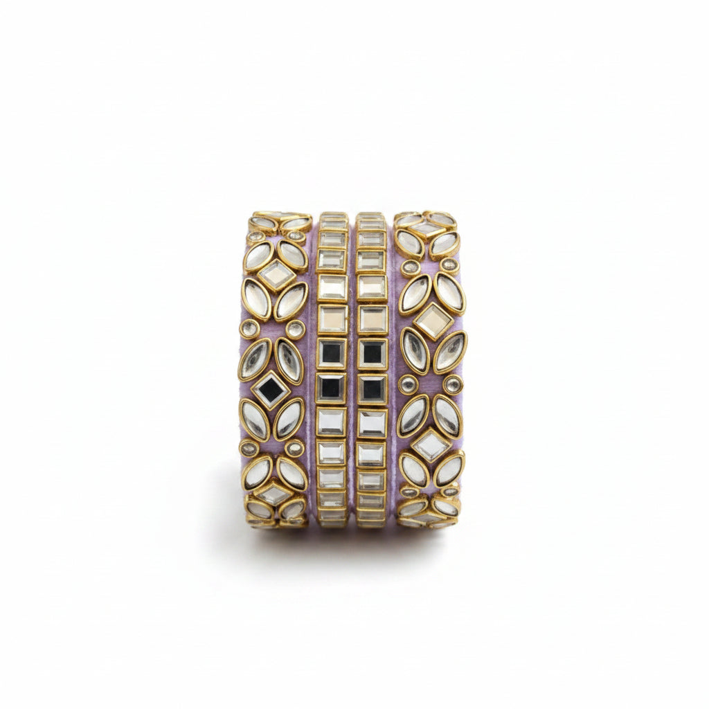 A wide gold-toned cuff bracelet embellished with floral inlay and clear stones, styled on a cream fabric background with a draped lavender scarf and small green eucalyptus sprigs.