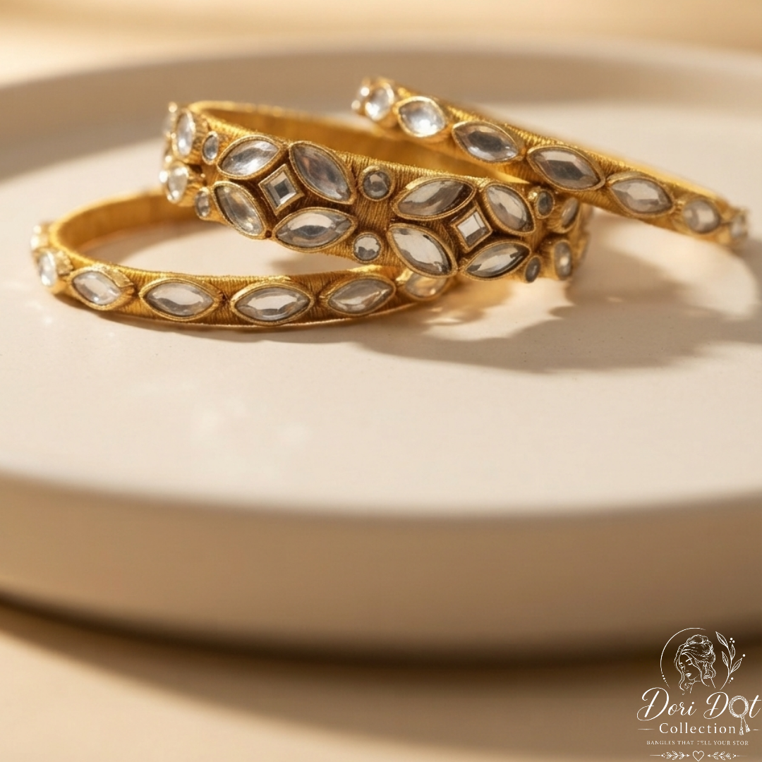 Three gold bangles adorned with clear teardrop-shaped stones, arranged on a round beige plate against a warm beige background. A 'Dose Dot Collection' logo is visible in the bottom right corner.