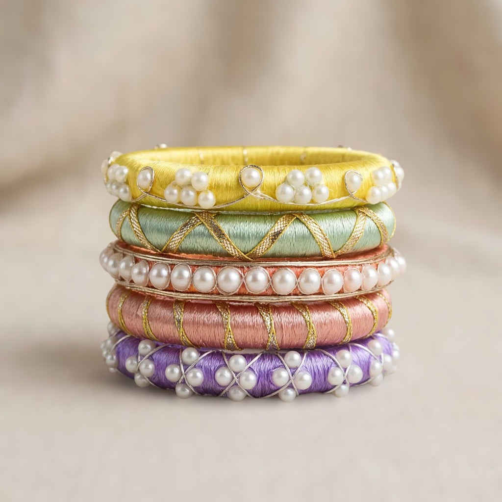A stack of five traditional Indian bangles (kara chandbali) made of vibrant silk threads in yellow, mint green, rose gold, pink, and purple, each adorned with white faux pearls and gold wire detailing, set against a soft beige background.