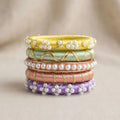 A stack of five traditional Indian bangles (kara chandbali) made of vibrant silk threads in yellow, mint green, rose gold, pink, and purple, each adorned with white faux pearls and gold wire detailing, set against a soft beige background.