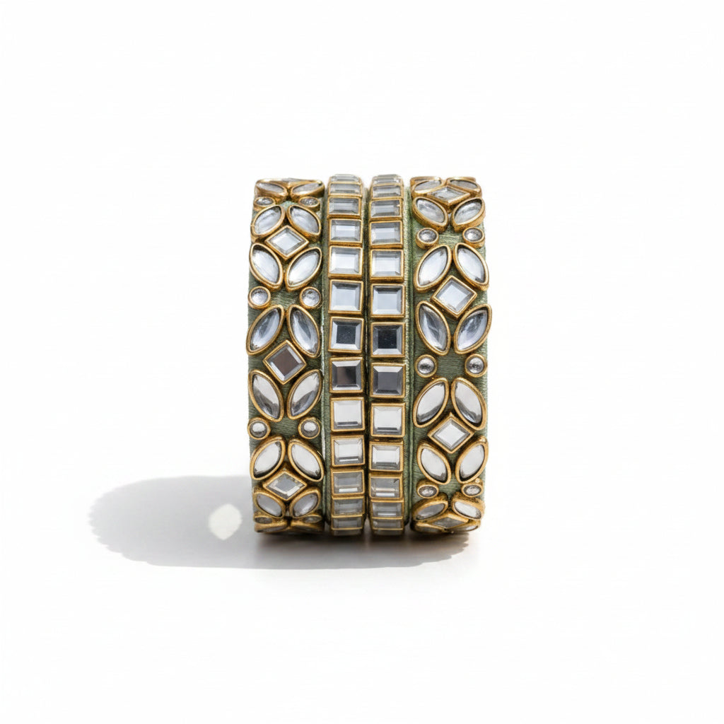A wide gold-toned cuff bracelet embellished with multiple rows of clear mirror work and white stones, styled on a cream embroidered fabric with sage green draped fabric and small white flowers.
