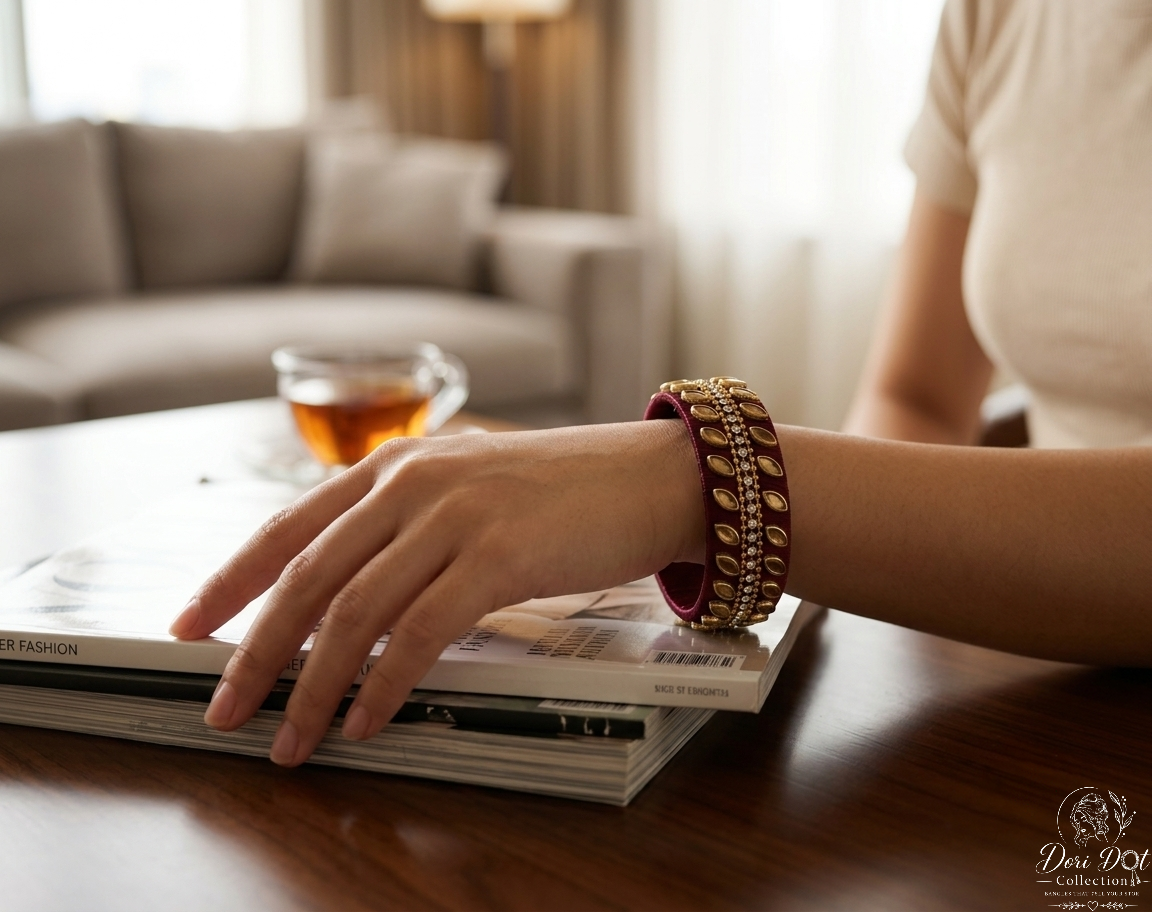 A close-up of a person's hand resting on a stack of magazines on a wooden table, wearing a stack of three gold and red bangles (kara) with a matching red bangle underneath. A glass of tea is visible in the soft-focus background of a living room setting.