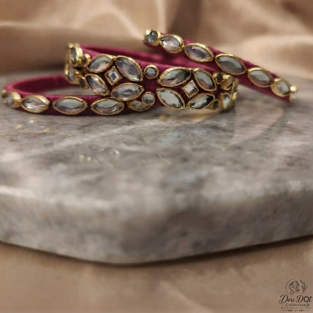 A pair of gold-plated bangles decorated with marquise and oval silver-toned stones, layered over a deep magenta velvet fabric, displayed on a gray marble slab with a beige satin background.