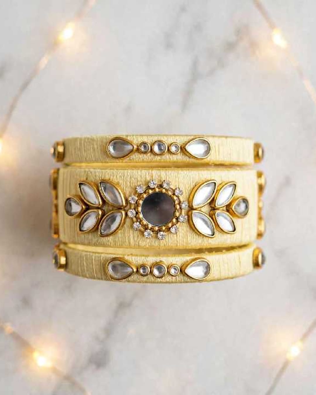A wide gold-toned stack cuff bracelet with intricate silver-toned stone and crystal embellishments, displayed on a light marble surface with warm bokeh string lights in the background.