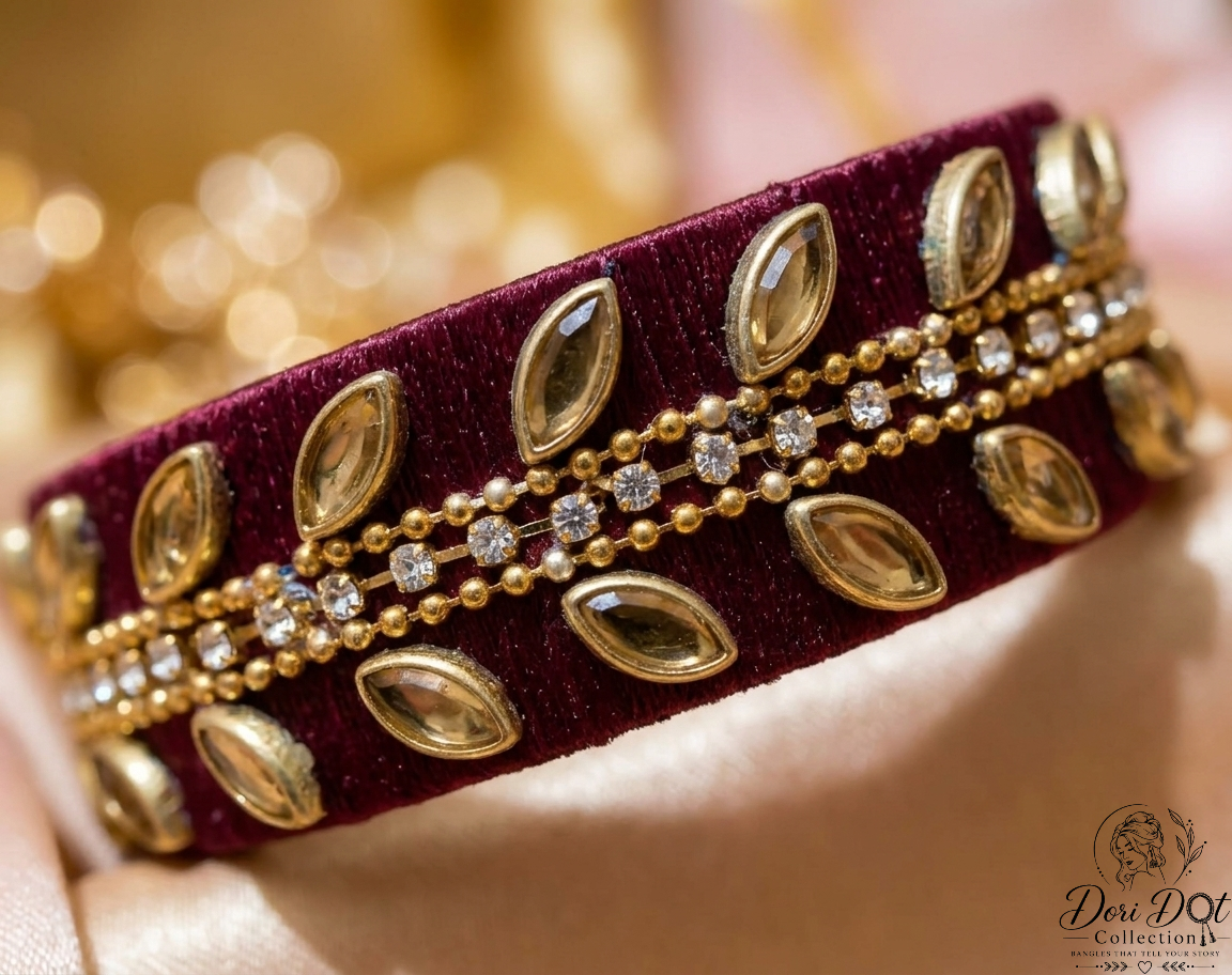 A close-up of a wide velvet bracelet in deep burgundy with ornate gold-toned beadwork, marquise-shaped metallic embellishments, and clear rhinestone accents, resting on soft cream fabric.