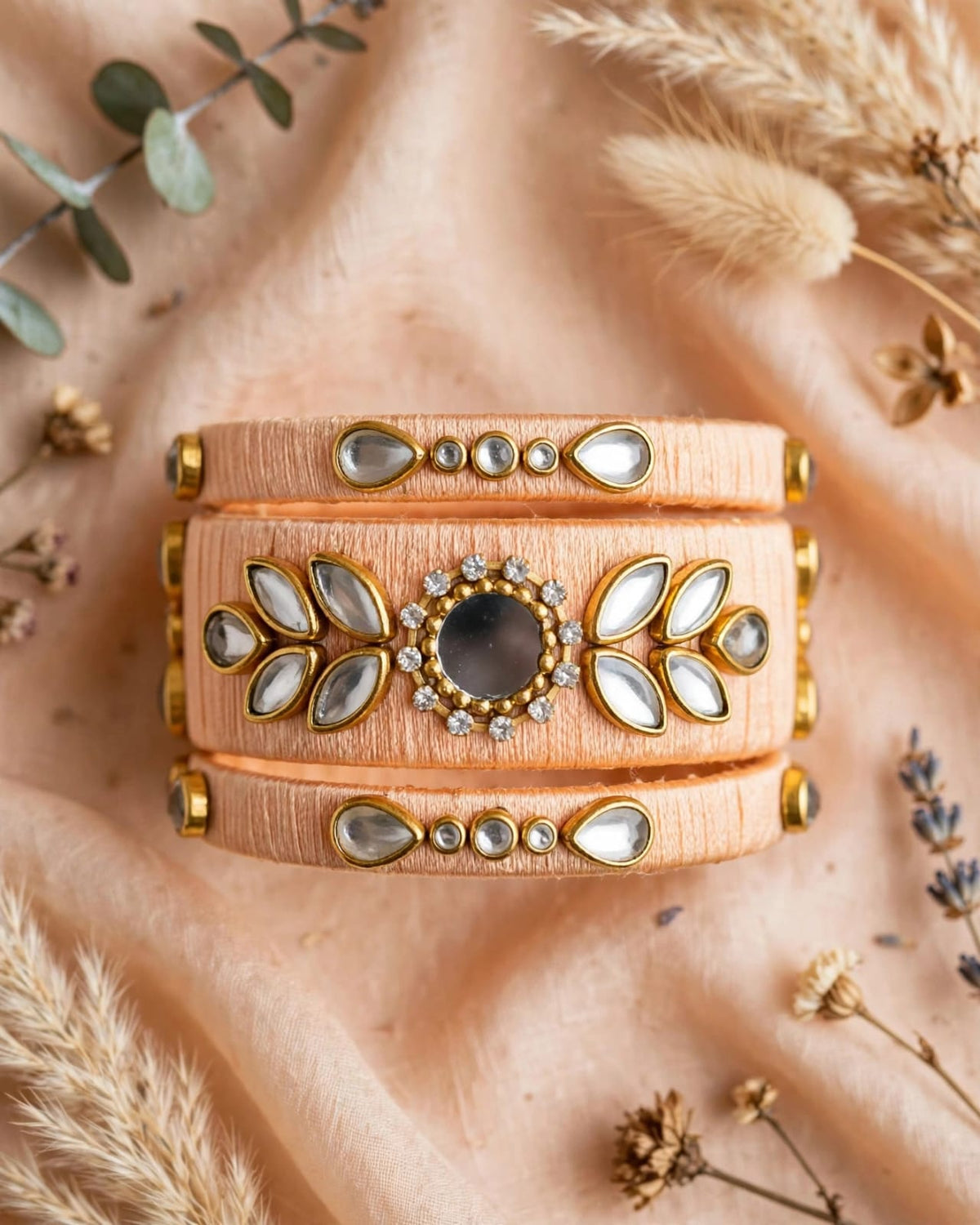 A wide three-band cuff bracelet in peach-colored textured leather with gold-tone metal accents, clear teardrop rhinestones, and a central round dark stone, styled on a peach fabric background with dried grasses and eucalyptus.