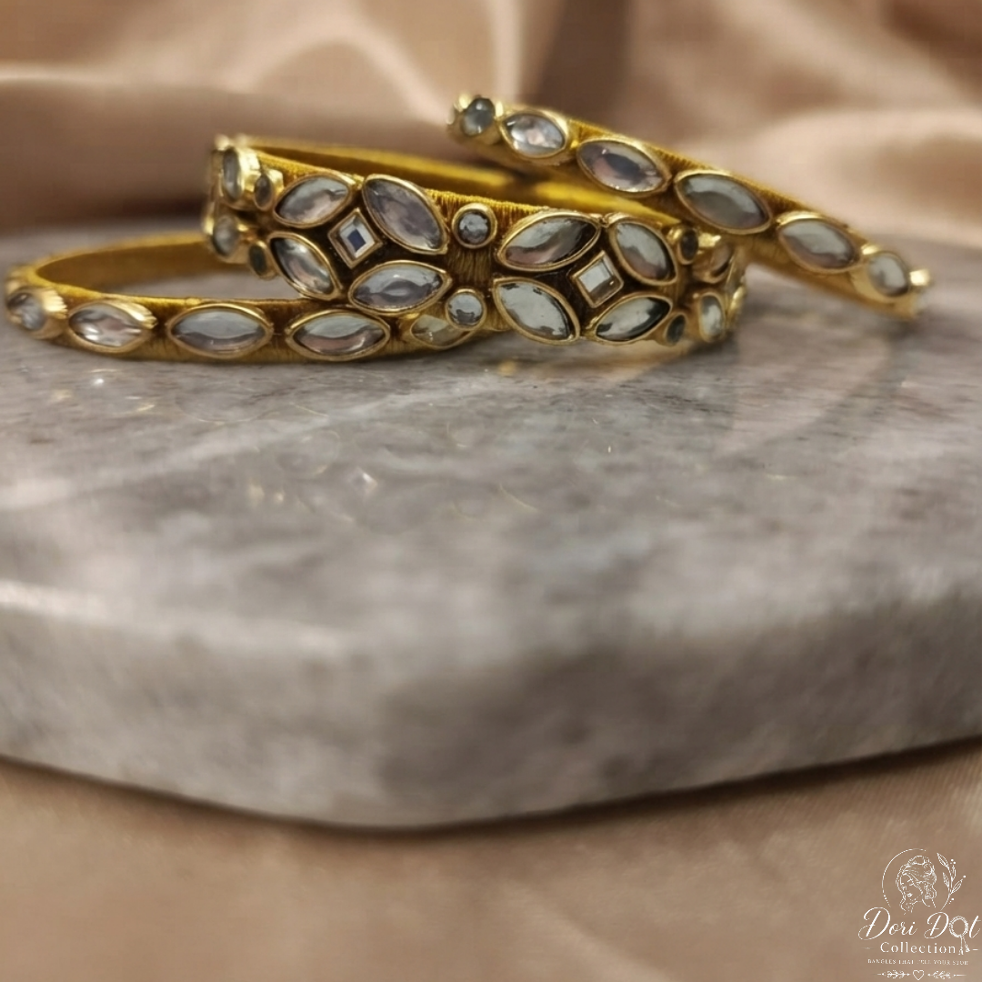 A pair of gold-plated bangle bracelets decorated with inlaid white stones, displayed on a gray marble slab with a beige satin fabric background.