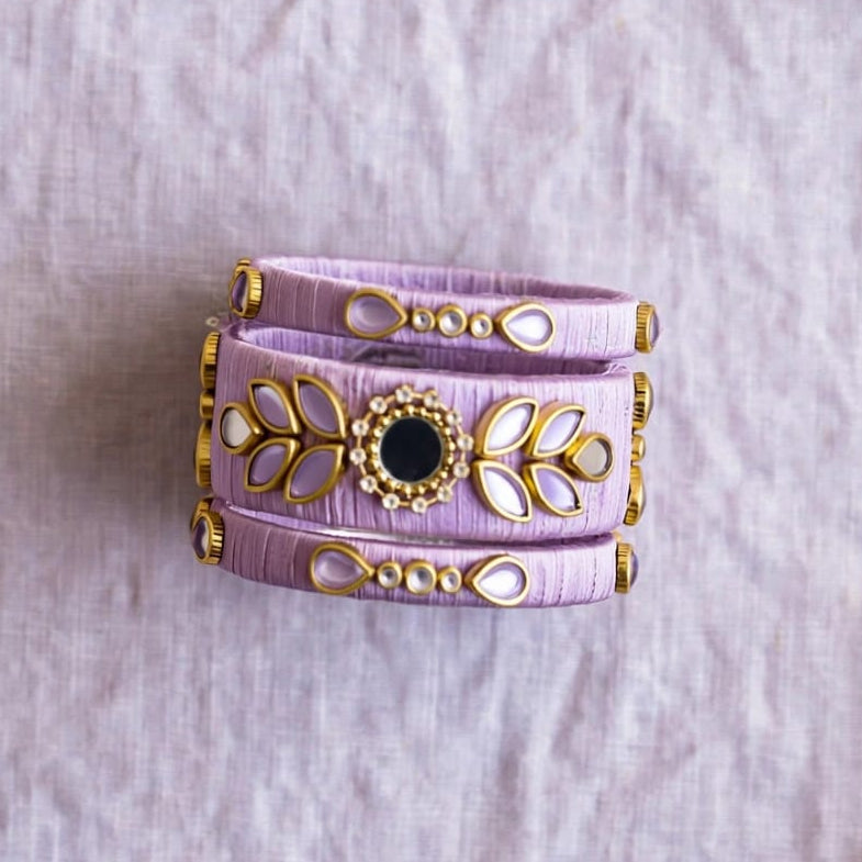 A lavender wrap bracelet with gold-toned filigree and a central black stone, styled on a textured lavender fabric with dried grass and baby's breath flowers.