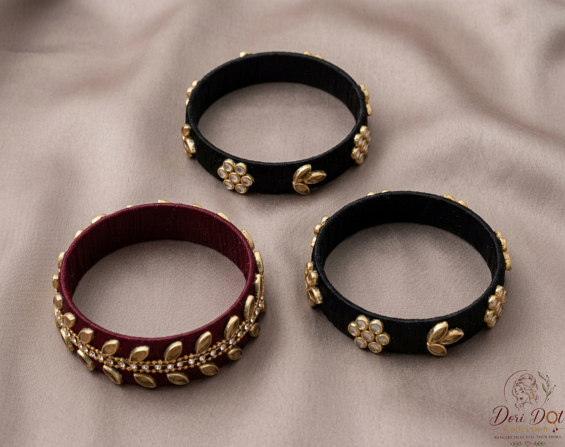 Three black leather bangles with gold floral and leaf embellishments, displayed on a beige fabric background. One bangle is shown from the outside, revealing a black exterior and gold leaf details.