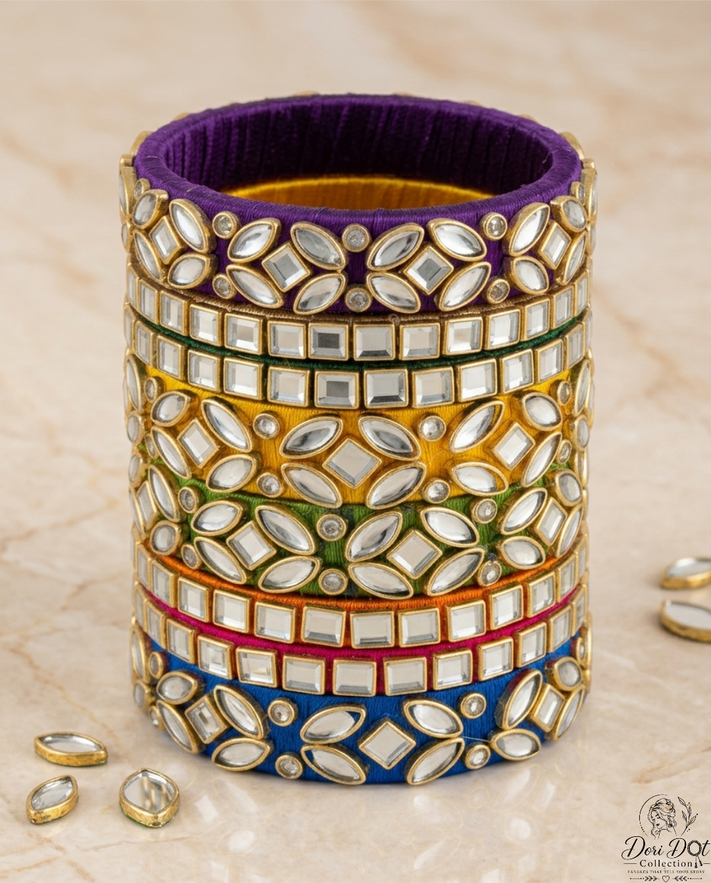 A stack of five traditional Indian bangles (kara) with silver mirror work (chikankari) and gold-plated borders, featuring purple, yellow, green, pink, and blue velvet fabric bands. The bangles are displayed on a beige marble surface with a few loose mirror stones in the foreground.