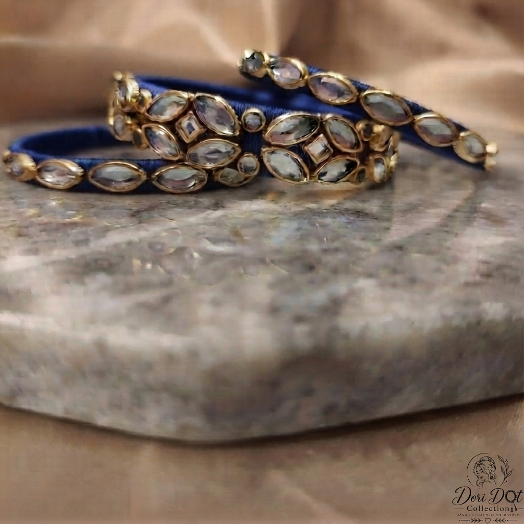 Three blue velvet bangles with gold-plated settings and clear stones, arranged on a gray stone slab with a beige fabric background.