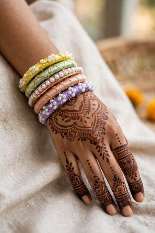 A close-up of a person's hand with intricate brown henna designs, adorned with a stack of colorful embroidered bangles featuring pearl-like beads, resting on a draped beige fabric.