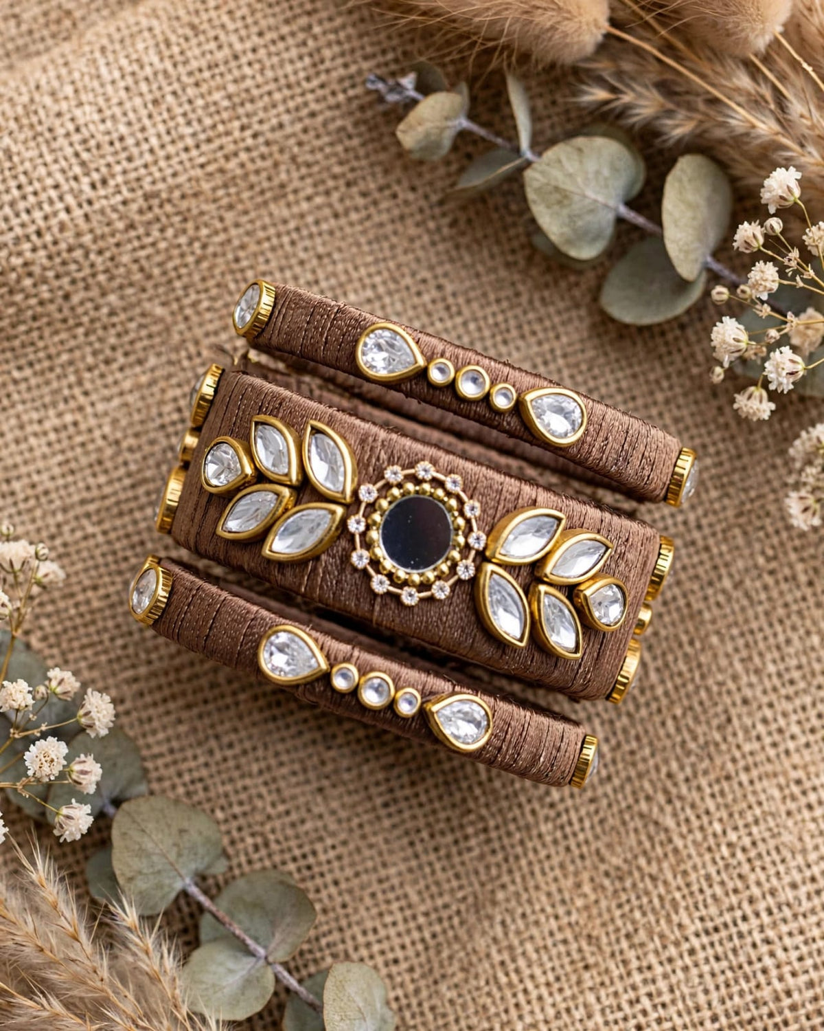 A wide brown fabric cuff bracelet with gold-tone metal trim, clear rhinestone accents, and a central dark oval stone, styled on a burlap surface with dried grass and eucalyptus leaves.