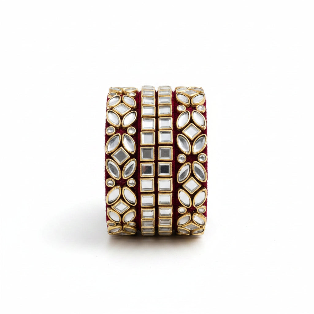 A wide gold-toned bangle bracelet decorated with multiple rows of white stones and a deep red velvet interior, styled on a white marble surface with draped white fabric and gold leaf accents.