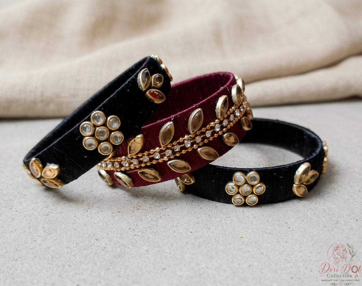 Three velvet bangles in black and deep red, decorated with gold leaf-shaped metalwork and clear stones, arranged on a light gray surface with a beige fabric background.