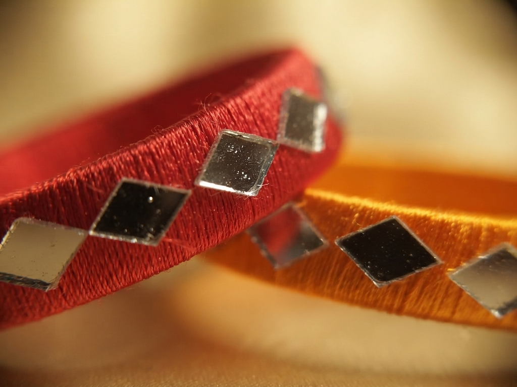 A close-up of two woven fabric bands, one red and one orange, each decorated with small square silver mirror-like accents, set against a warm blurred background.