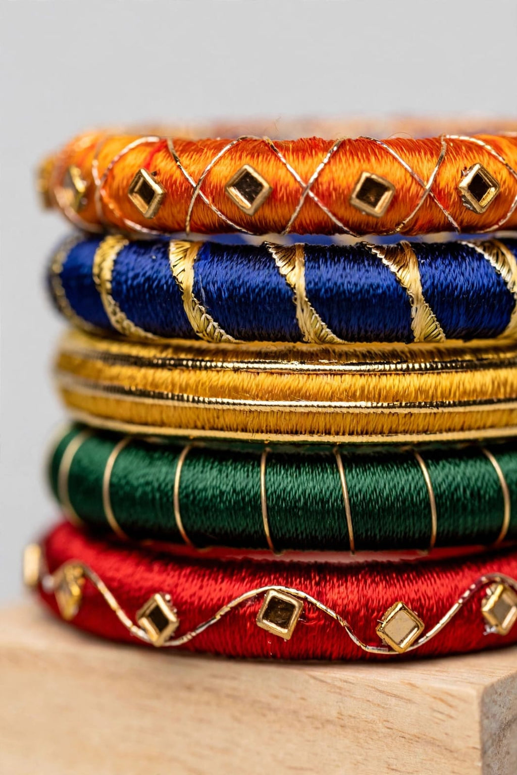 A stack of five traditional Indian silk bangles in orange, blue, gold, green, and red, each decorated with gold thread patterns and small metallic diamond-shaped studs, displayed on a light wooden surface.