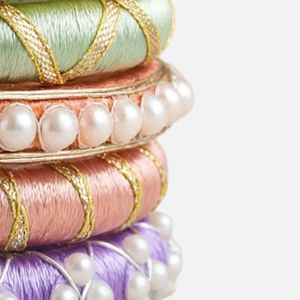 A close-up stack of decorative bangles or rings made from stacked silk ribbons in pastel green, peach, pink, and lavender, each accented with gold braided trim and white faux pearls, set against a plain light gray background.