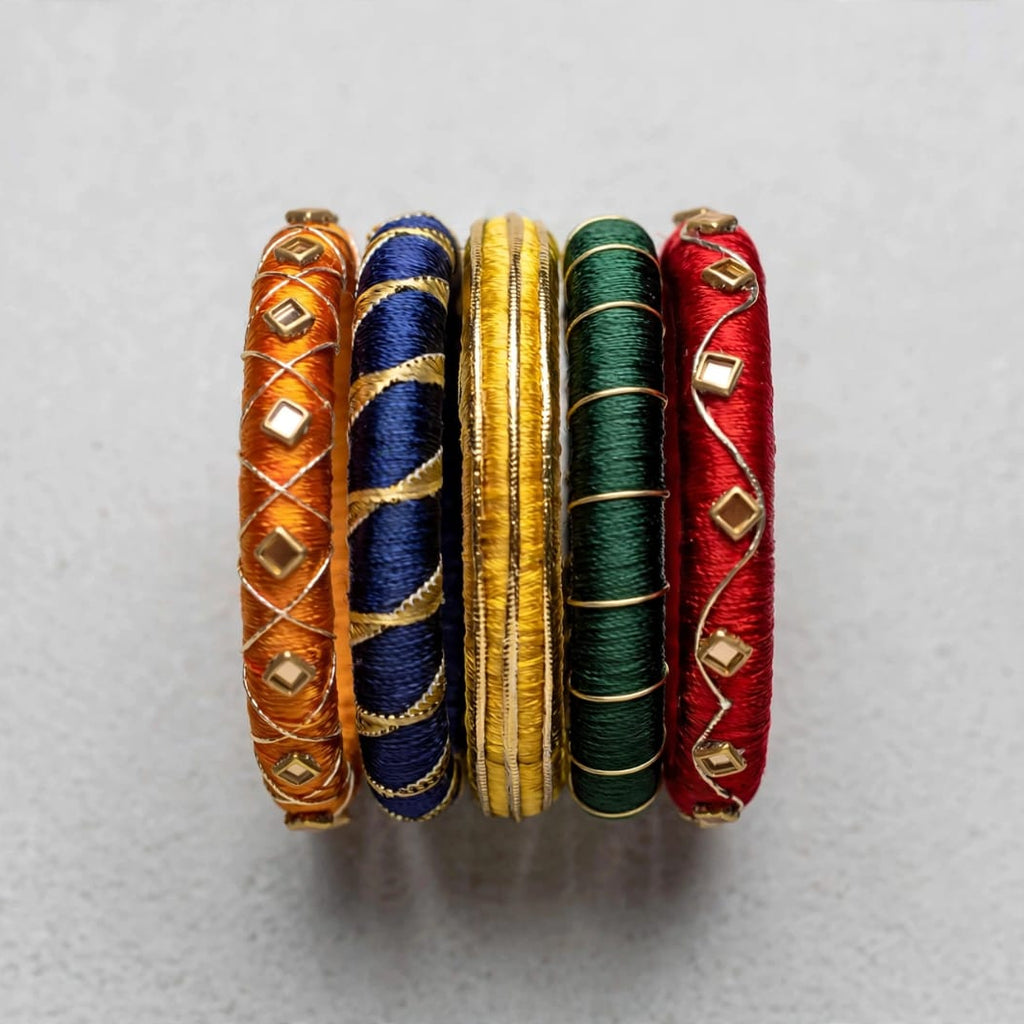 Four traditional Indian bangles made of wound silk threads in orange, navy blue, gold, and green, embellished with small gold diamond-shaped beads, arranged vertically on a light gray background.