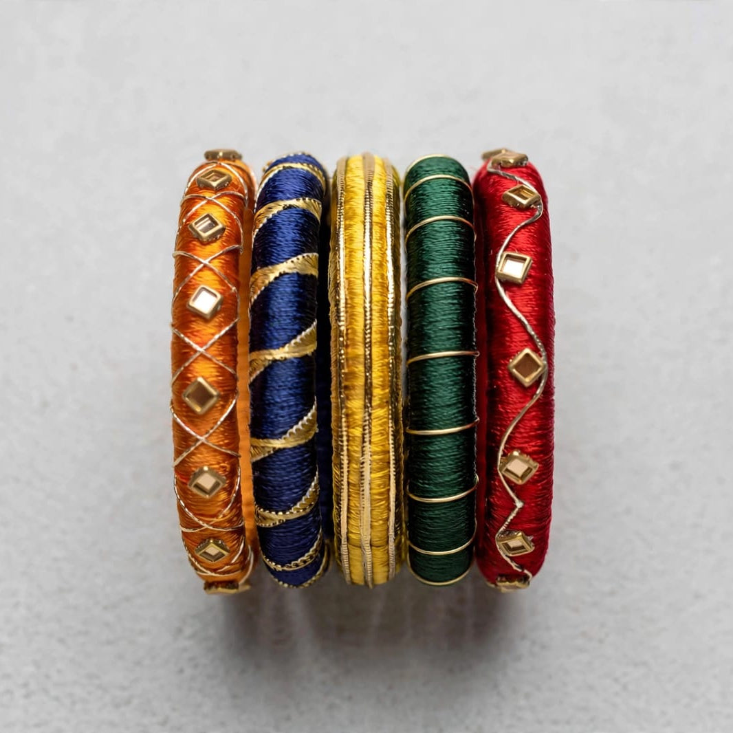Four traditional Indian bangles made of wound silk threads in orange, navy blue, gold, and green, embellished with small gold diamond-shaped beads, arranged vertically on a light gray background.