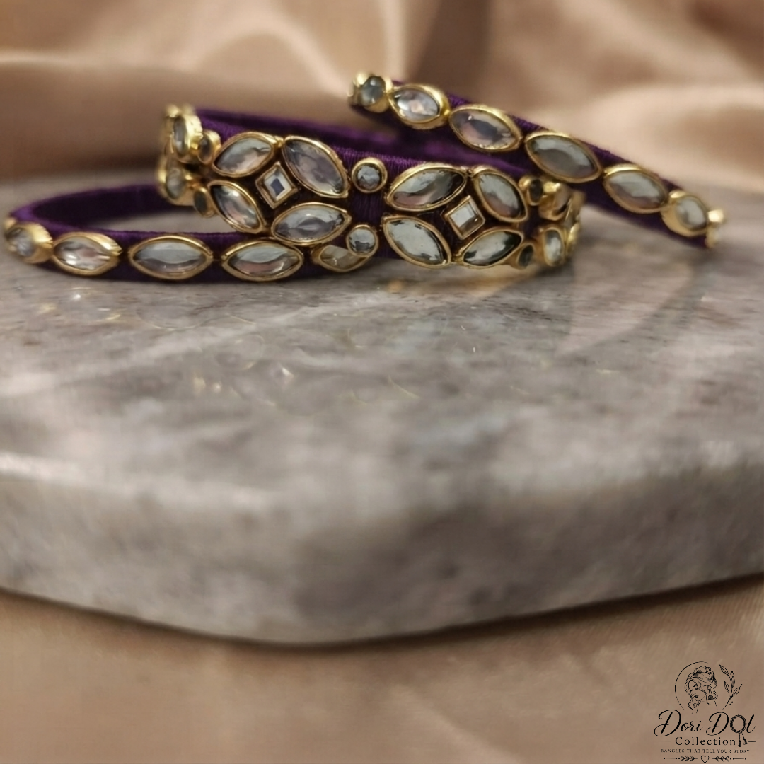 A close-up of a purple velvet bangle bracelet with gold-plated settings and inlay of clear stones, resting on a gray marble slab with a beige satin fabric background.