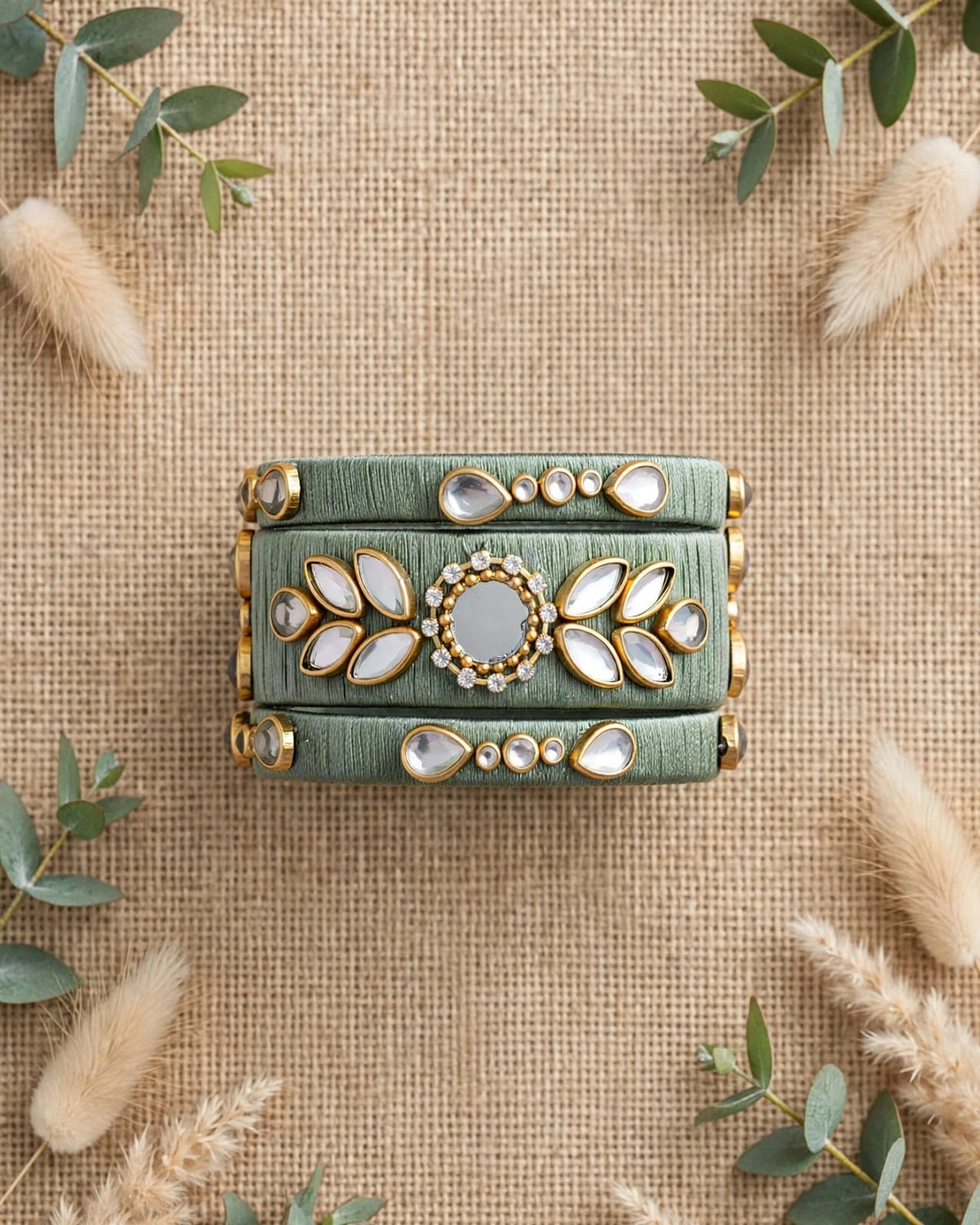 A rectangular sage green clutch bag with gold-tone hardware and white stone embellishments, styled flat-lay on a burlap background with decorative greenery.
