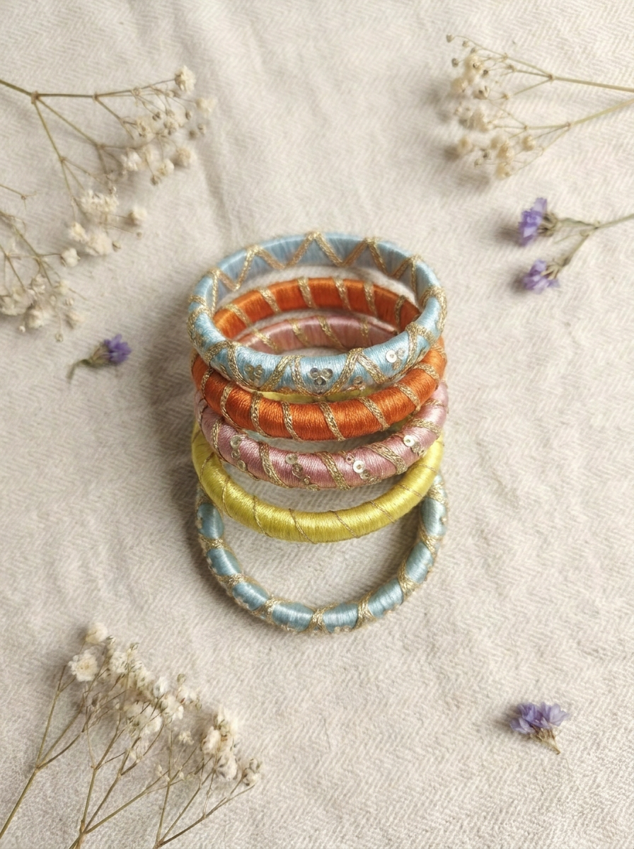 A stack of five patterned bangles in light blue, orange, pink, and yellow with gold foil detailing, arranged on a textured beige fabric with dried white and purple flowers.