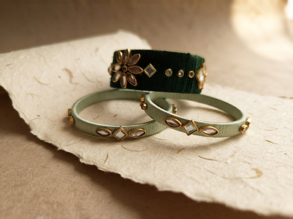 Three decorative bangles displayed on a piece of textured beige paper: two pale sage green bangles in the foreground and one dark forest green bangle in the background, both adorned with gold-toned floral and diamond-shaped embellishments.