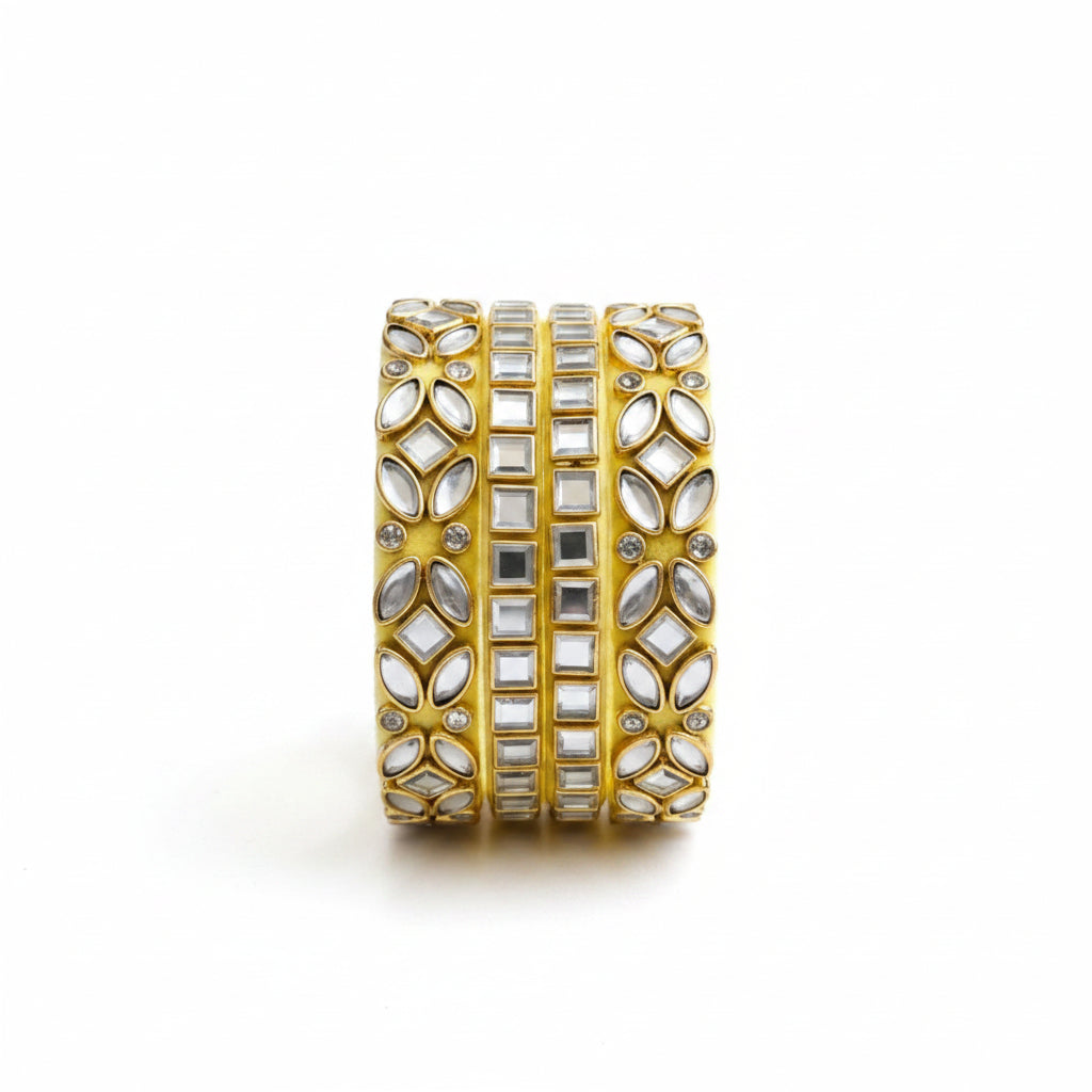 A wide gold cuff bracelet embellished with multiple rows of clear and silver mirror-polished stones, styled on a light fabric background with draped pale yellow fabric and small white baby's breath flowers.
