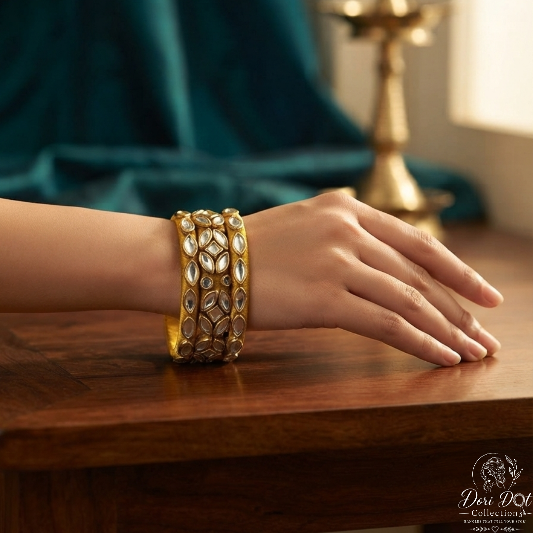 A close-up of a person's hand wearing a wide gold bangle with intricate silver and white stone embellishments, resting on a wooden table with a dark teal drape and a brass lamp in the background.